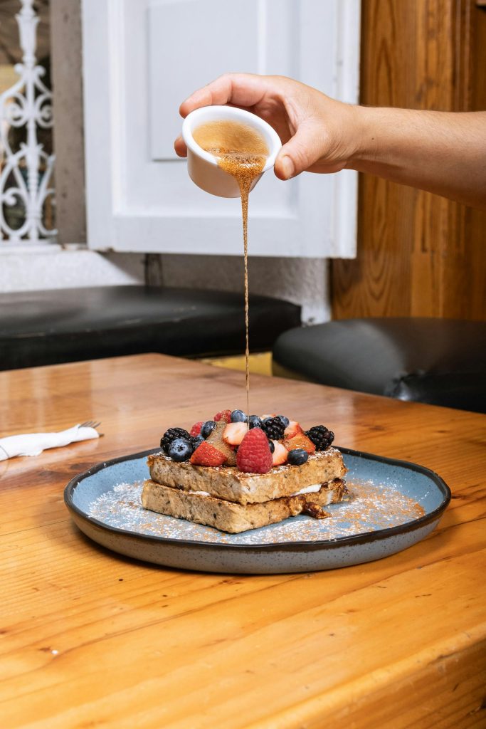 French toast topped with fresh berries and syrup pouring from a dish, set on a wooden table.
