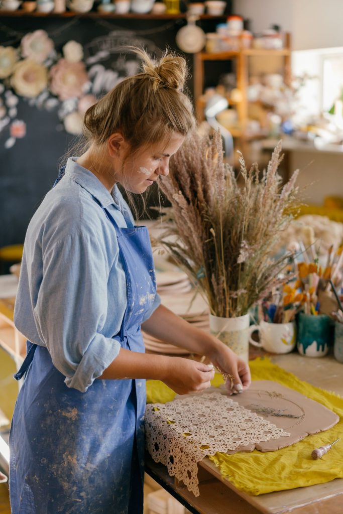 A woman artist in a blue apron crafts clay art at a sunlit workshop table, surrounded by tools and dried flowers.