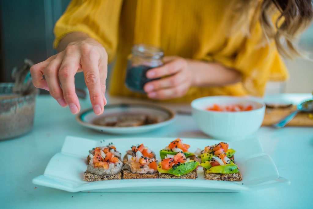 Hand sprinkling seeds on avocado toast with diced tomatoes, featuring a fresh and healthy dining setup.